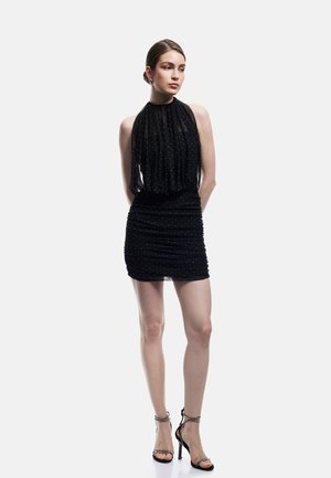 Giovane donna con mini abito nero smanicato a pois piccoli, indossa sandali neri con tacco alto, in piedi davanti a uno sfondo semplice.