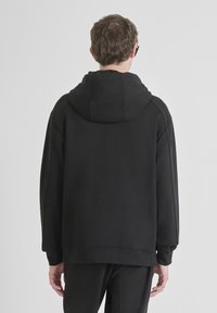 Hombre vistiendo un sudadera con capucha negra y pantalones negros, de pie frente a una pared gris claro lisa con las manos relajadas a los lados.
