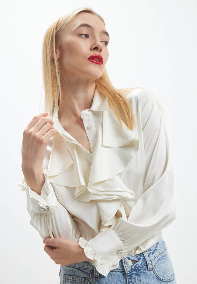 Blonde woman with red lipstick wearing a white ruffled blouse and blue jeans, posing with one hand near her face and looking sideways.