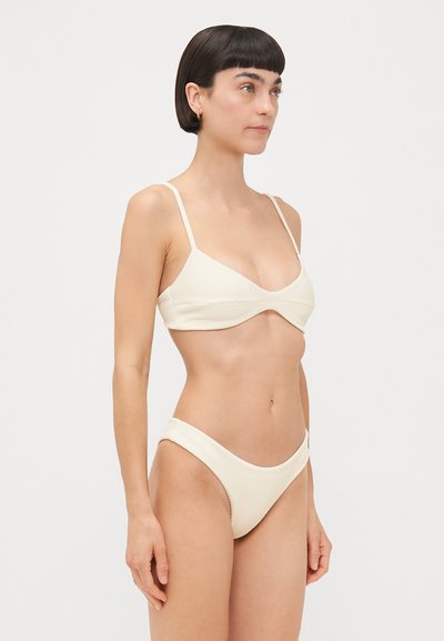 Krēmkrāsas ribota bikini komplekts, kuram ir braletu stila augšdaļa ar plānām siksniņām un vidējas seguma apakšdaļa ar zemu jostasvietu.