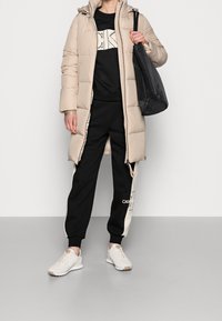 Abrigo acolchado beige con capucha, sudadera negra, pantalones de chándal negros con acentos blancos y zapatillas ligeras. Bolsa de mano negra.