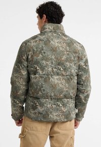 Giubbotto imbottito con un motivo camouflage nei toni del verde, marrone e grigio. Collare alto e dettagli cuciti sono ben visibili.