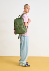 Mochila de nailon verde con un bolsillo delantero con cremalleras, correas ajustables y una etiqueta de llavero. Llevada por una persona con una camiseta rosa y jeans claros.