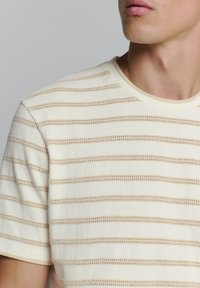 Helles Creme-T-Shirt mit horizontalen beigen Streifen, das ein strukturiertes Muster und einen Rundhalsausschnitt aufweist. Weicher Stoff mit lockerer Passform.