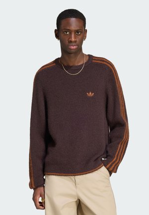 Joven con un suéter marrón oscuro con rayas naranjas en las mangas y pantalones beige, de pie con una mano en el bolsillo.