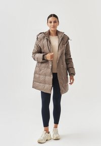 Bruine pufferjas met een capuchon, drukknoopsluiting en een geborduurd ontwerp. Draagt over een beige top, gecombineerd met marineblauwe leggings en lichtgekleurde sneakers.