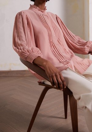 Blouse rose avec plis, fermeture à boutons et col à volants. Tissu transparent avec manches longues froncées, portée par un mannequin assis.