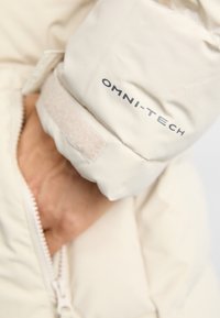 Sildītājs gaiši iedeguma krāsā ar tuvplānu pie manšetes, kurā ir velcro josla un "OMNI-TECH" logo pelēkā krāsā. Gluda, izolēta auduma.