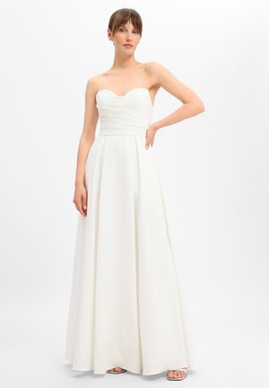 Vrouw draagt een strapless, witte, lange jurk met een aansluitend lijfje en een volle rok, staand met één hand op haar heup.