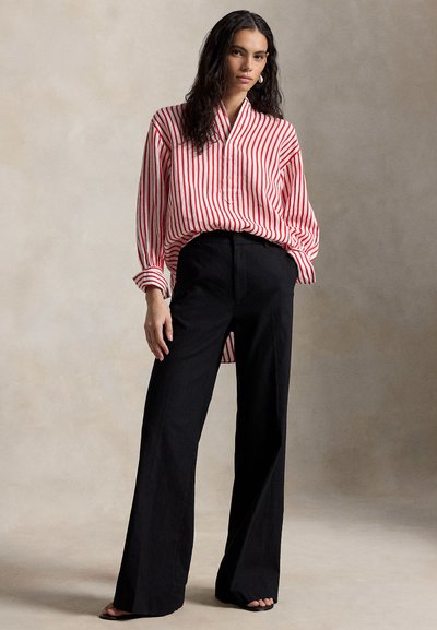 Osoba oblečená v červeno-bielej zvislo pruhovanej voľnej blúzke, ktorá je zastrčená do čiernych širokých nohavíc s vysokým pásom, stojaca pred jednoduchým pozadím.