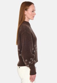 Brun ullgenser med høy hals, nedfalne skuldre og hvite "H" mønstre på ermene. Teksturert strikk og ribbede mansjetter.