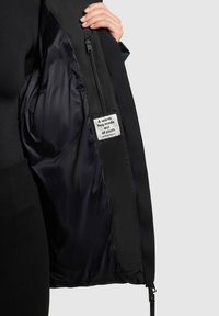 Crna jakna s glatkom, svilenom unutrašnjošću. Ima džep s patentnim zatvaračem i oznaku koja kaže: "Topli zagrljaj napravljen od tkanine."