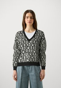 Maglione grigio con un motivo geometrico nero, con scollatura a V, maniche lunghe e polsini a costine. Indossato sopra una maglietta bianca, abbinato a pantaloni in denim chiaro.