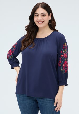 Uśmiechnięta kobieta z długimi ciemnymi włosami ubrana w granatową bluzkę z kolorowym kwiatowym haftem na rękawach i niebieskie dżinsy.