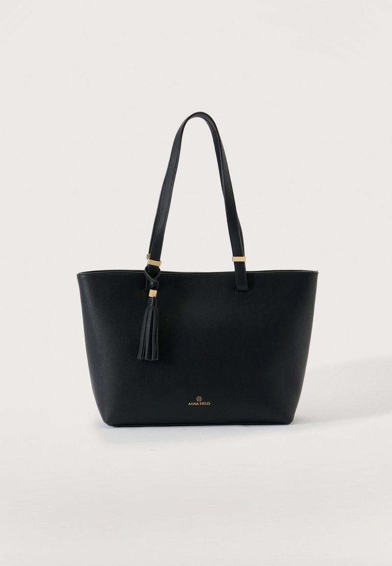 Sac cabas noir avec deux longues poignées et un détail pompon, petit logo doré "Anna Field" au centre avant sur fond uni.