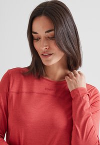Woman with medium-length dark hair wearing a long-sleeve red shirt with "TOG24" logo, looking down and touching her neck.