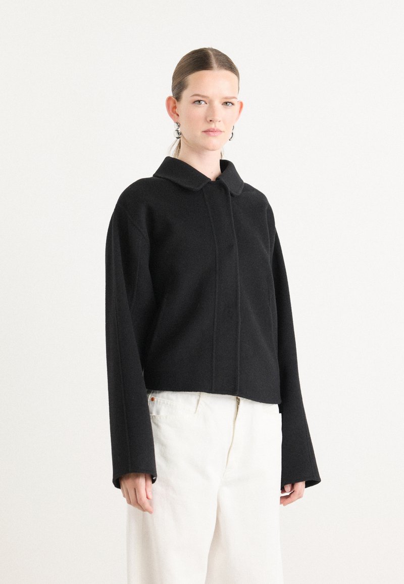 Zwarte cropped jas met een ronde kraag, lange mouwen en een textuurafwerking. Gecombineerd met crèmekleurige broeken, heeft het een minimalistisch ontwerp.