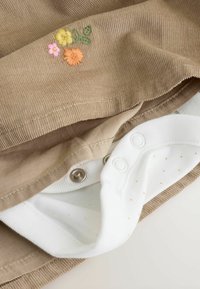 Kordon kledingstuk in beige met bloemenborduurwerk in roze, geel en oranje. Binnenlaag is wit met kleine oranje stippen en drukknoopjes.