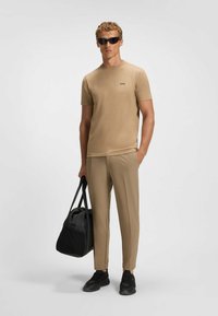 Beige Kurzarm-T-Shirt mit schwarzem Logo, kombiniert mit hellbraunen, maßgeschneiderten Hosen. Er trägt eine schwarze Tasche und schwarze Sneakers.