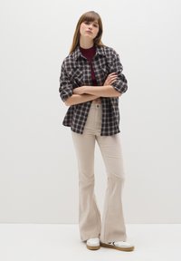 Camisa de franela a cuadros en azul marino y beige, combinada con pantalones de campana beige y zapatillas blancas. El modelo está de pie con los brazos cruzados frente a un fondo liso.
