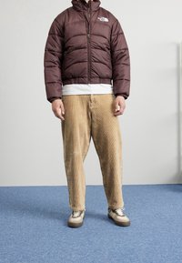 Kurtka puchowa w ciemnym fioletowym kolorze z zamkiem błyskawicznym, prążkowanymi mankietami oraz tonowym logo. Połączona z beżowymi spodniami z kordurowej tkaniny i trampkami z beżowymi akcentami.