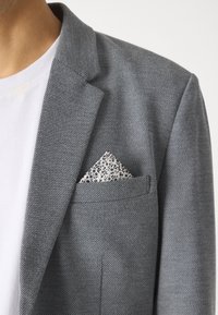 Grå blazer med en strukturerad väv, som har en vit T-shirt under och en vikt vit näsduk med svart blommönster.