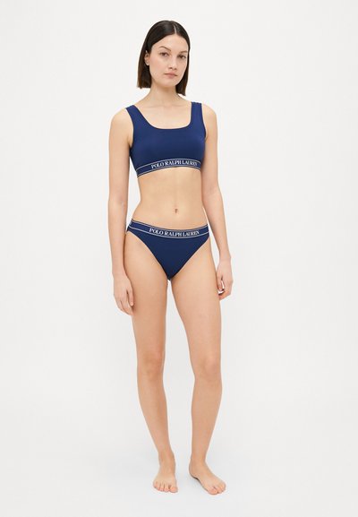 Tamsiai mėlynas bralette ir bikini rinkinys, su balta ir mėlyna logo juosta. Lygus audinys su prigludusia, šiuolaikiška dizainu.