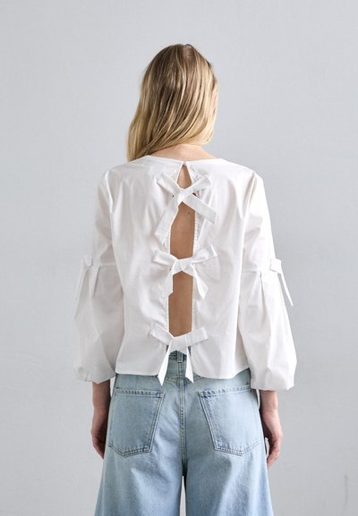 Fehér pamut blúz hosszú ujjakkal, hátul kivágással és több szövetmasnival. Világoskék, bőszárú farmerral párosítva.