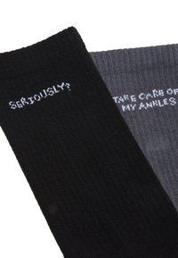 Schwarze und graue Socken mit gerippter Textur. Die schwarze Socke hat den Text "ERNSTHAFT?" und die graue Socke trägt "KÜMMERE DICH UM MEINE KNÖCHEL", beides in Weiß.