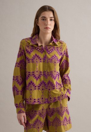 Junge Frau mit langen Haaren trägt ein olivgrünes, knopfgelegenes Hemd und passende Shorts mit lila, filigranen Blumenmustern, stehend vor einfarbigem Hintergrund.