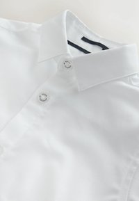 Baltas marškinių apykaklės ir viršutinių sagų artimas vaizdas, ant sagų užrašyta „Baker by Ted Baker“.