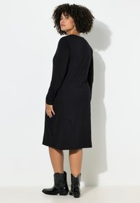 Zwarte jurk met lange mouwen en een losse pasvorm, reikt tot halverwege de kuit, gecombineerd met zwarte enkellaarzen met zilverkleurige accenten.