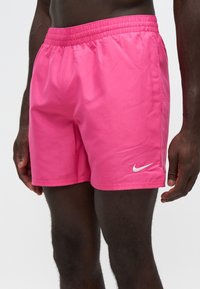 Άνδρας που φοράει φωτεινά ροζ σορτς Nike με ελαστική μέση και λευκό λογότυπο Nike swoosh στο αριστερό πόδι σε απλό φόντο.
