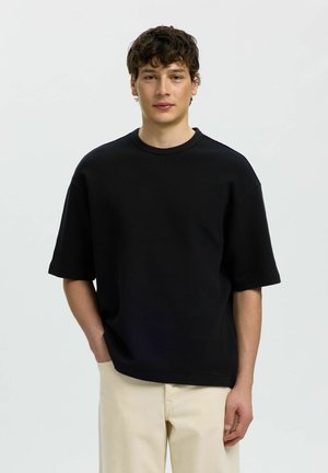 Jeune homme aux cheveux courts et bruns porte une chemise noire ample à manches courtes et un pantalon beige clair, debout devant un fond clair uni.