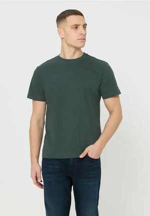 Giovane uomo che indossa una maglietta verde scuro a maniche corte e jeans blu scuro, in piedi con una mano in tasca, guardando di lato.