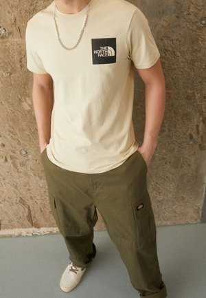 Mann trägt ein beiges The North Face T-Shirt, olivgrüne Cargohosen, weiße Turnschuhe und eine silberne Kettenhalskette vor einer strukturierten Wand.