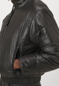 Svart vadderad jacka med hög krage, glansigt material, elastiska muddar, fickor med dragkedja och kort design. Synligt texturerat ytskikt.