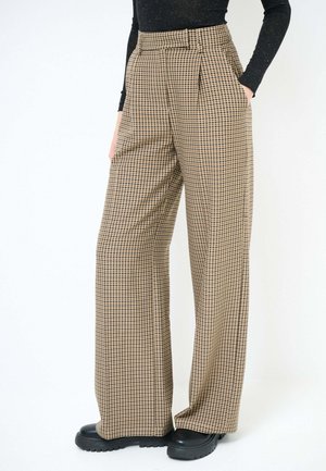 Persona che indossa pantaloni marroni a vita alta a quadri con gamba larga, scarpe nere con suola spessa e un top nero a maniche lunghe.