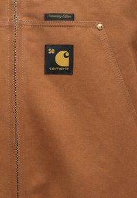 Καφέ καμβάς ζακέτα με χρυσό φερμουάρ, μαύρη αναμνηστική ετικέτα με τον αριθμό 50 και ετικέτα "Έκδοση Επετείου".