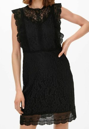 Robe noire sans manches en dentelle avec bords festonnés, col montant et motifs floraux complexes portée par une femme la main sur la hanche.