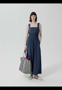 Woman in sleeveless navy jumpsuit holding large black-and-white striped tote with colorful trim, wearing beige woven slip-on shoes.