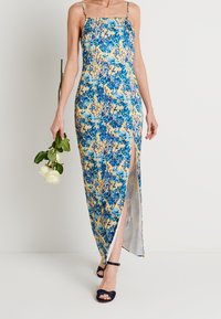 Blumenkleid im Maxistil mit blauen und gelben Mustern, figurbetonter Silhouette, seitlichem Schlitz und dünnen Spaghettiträgern. Die Schuhe sind navyfarbene Riemchenheels.