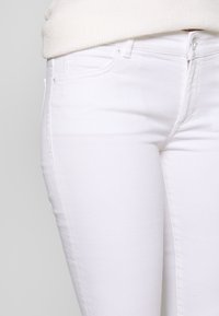 Bílé džínové kalhoty s přiléhavým střihem, vybavené předními kapsami, zapínáním na zip a hladkou texturou. Minimalistické švy jako ozdoba.