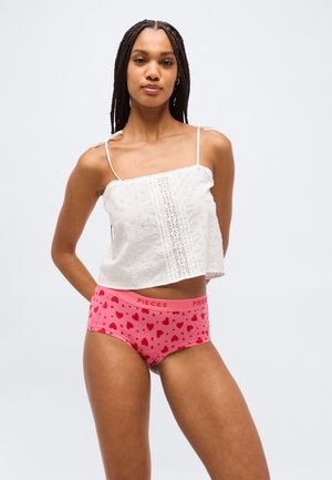 Γυναίκα με μακριά κοτσίδα που φοράει λευκό αμάνικο τοπ και ροζ εσώρουχο με σχέδιο καρδιές, στέκεται με τα χέρια πίσω από την πλάτη της.