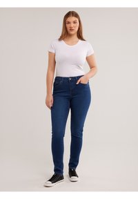 Sieviete stāv, valkājot baltu īsroku krekliņu, tumši zilas džinsas un melnas sporta kurpes, viena roka ielikta kabatā.