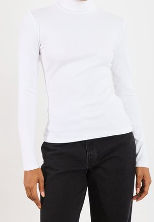 Personne portant un haut blanc moulant à col roulé et manches longues et un pantalon noir, debout devant un fond clair uni.