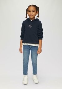 Ciemnoniebieska bluza z kapturem z napisem "Magia jest wszędzie", warstwowo założona na jasną koszulkę, w połączeniu z niebieskimi dżinsami i białymi trampkami.