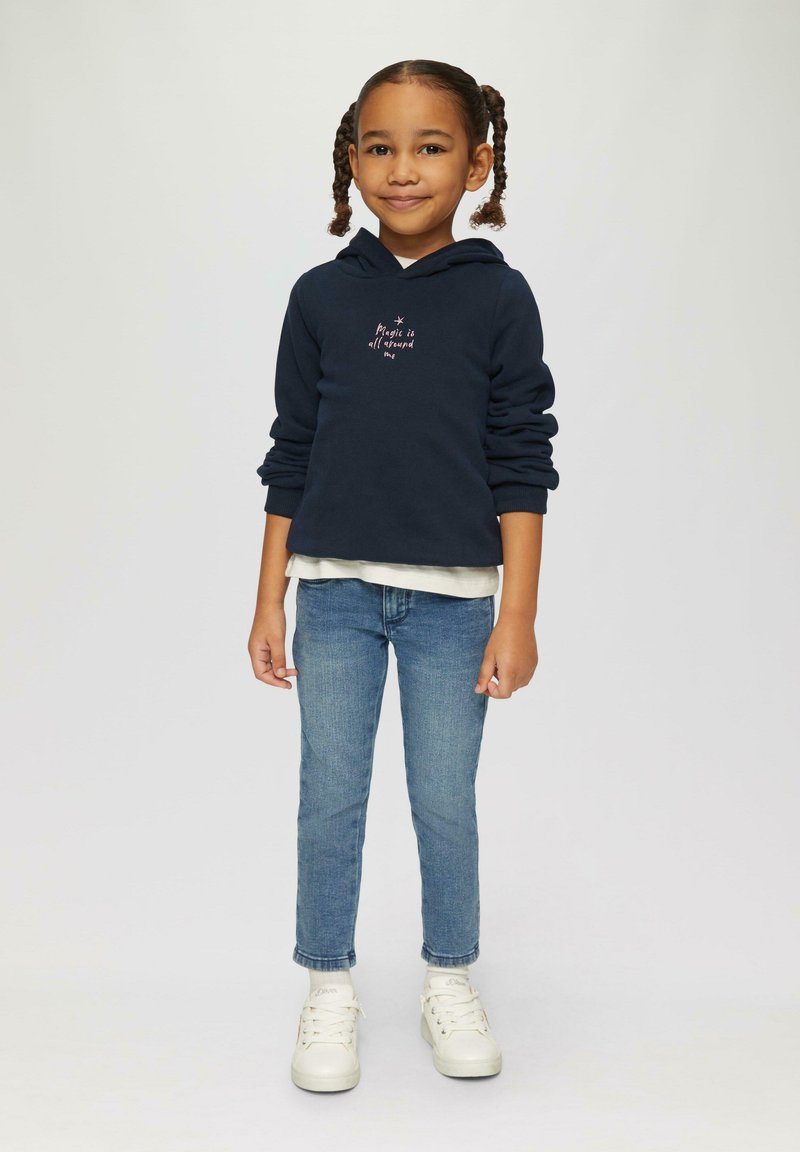 Ciemnoniebieska bluza z kapturem z napisem "Magia jest wszędzie", warstwowo założona na jasną koszulkę, w połączeniu z niebieskimi dżinsami i białymi trampkami.