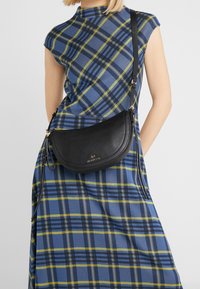 Bolso cruzado de cuero negro con forma curvada, herrajes dorados y un acabado texturizado. Lleva puesto sobre un vestido a cuadros en tonos azul y verde.