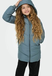 Light blue puffer jacket with a zip front, hood, and diagonal quilted pattern; worn over black pants, featuring long wavy hair framing the face.
